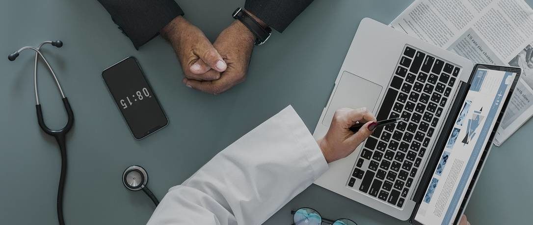 Profissional da saúde apresenta informações sobre plano de saúde empresarial a um gestor durante uma reunião, com notebook, tablet e estetoscópio sobre a mesa.