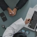 Profissional da saúde apresenta informações sobre plano de saúde empresarial a um gestor durante uma reunião, com notebook, tablet e estetoscópio sobre a mesa.