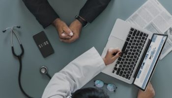 Profissional da saúde apresenta informações sobre plano de saúde empresarial a um gestor durante uma reunião, com notebook, tablet e estetoscópio sobre a mesa.
