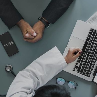 Profissional da saúde apresenta informações sobre plano de saúde empresarial a um gestor durante uma reunião, com notebook, tablet e estetoscópio sobre a mesa.