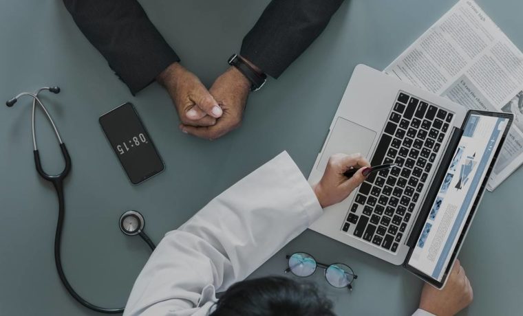 Profissional da saúde apresenta informações sobre plano de saúde empresarial a um gestor durante uma reunião, com notebook, tablet e estetoscópio sobre a mesa.