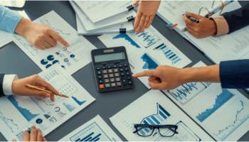 Pessoas analisando relatórios financeiros e gráficos ao redor de uma calculadora, representando o controle e o planejamento do fluxo de caixa de uma empresa.