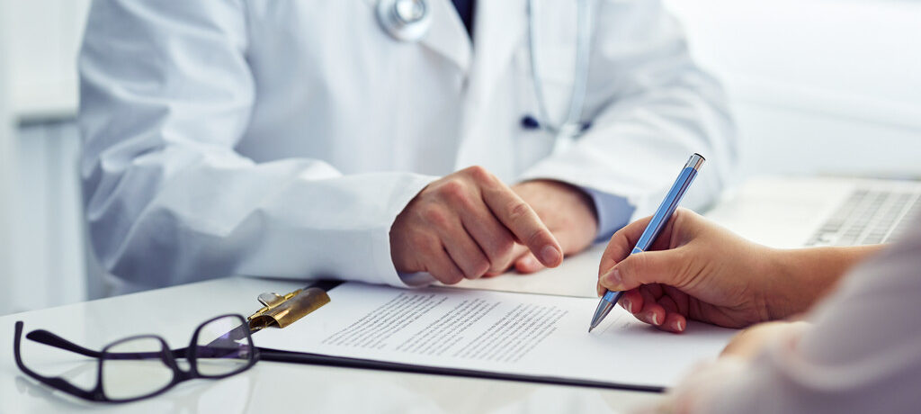 Médico de jaleco indica com o dedo onde o paciente deve assinar um documento de saúde sobre a mesa de atendimento.
