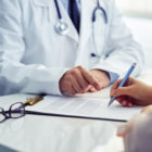Médico de jaleco indica com o dedo onde o paciente deve assinar um documento de saúde sobre a mesa de atendimento.