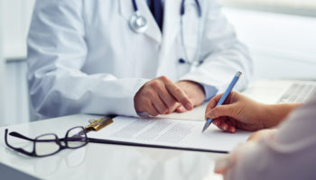 Médico de jaleco indica com o dedo onde o paciente deve assinar um documento de saúde sobre a mesa de atendimento.