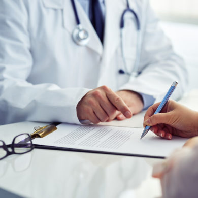 Médico de jaleco indica com o dedo onde o paciente deve assinar um documento de saúde sobre a mesa de atendimento.