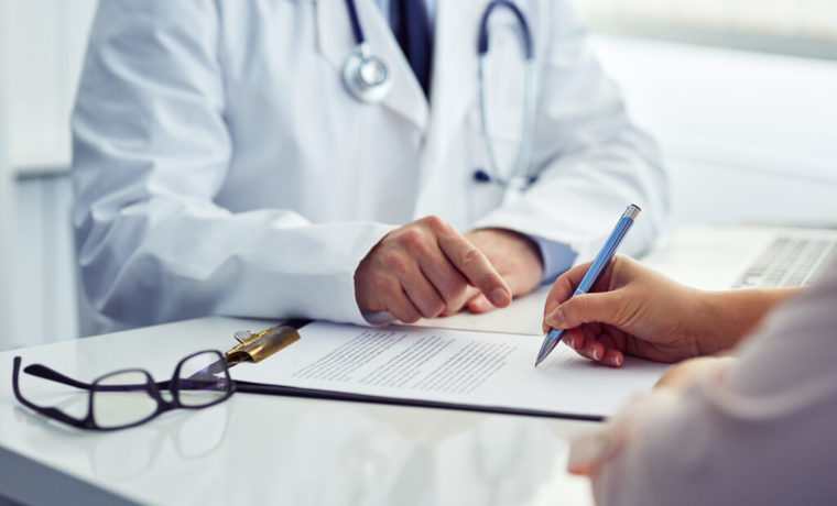 Médico de jaleco indica com o dedo onde o paciente deve assinar um documento de saúde sobre a mesa de atendimento.