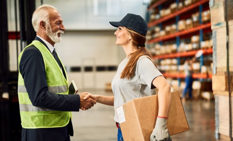 Fornecedor e profissional do setor logístico apertam as mãos em um centro de distribuição, simbolizando parceria comercial e negociação com fornecedores.
